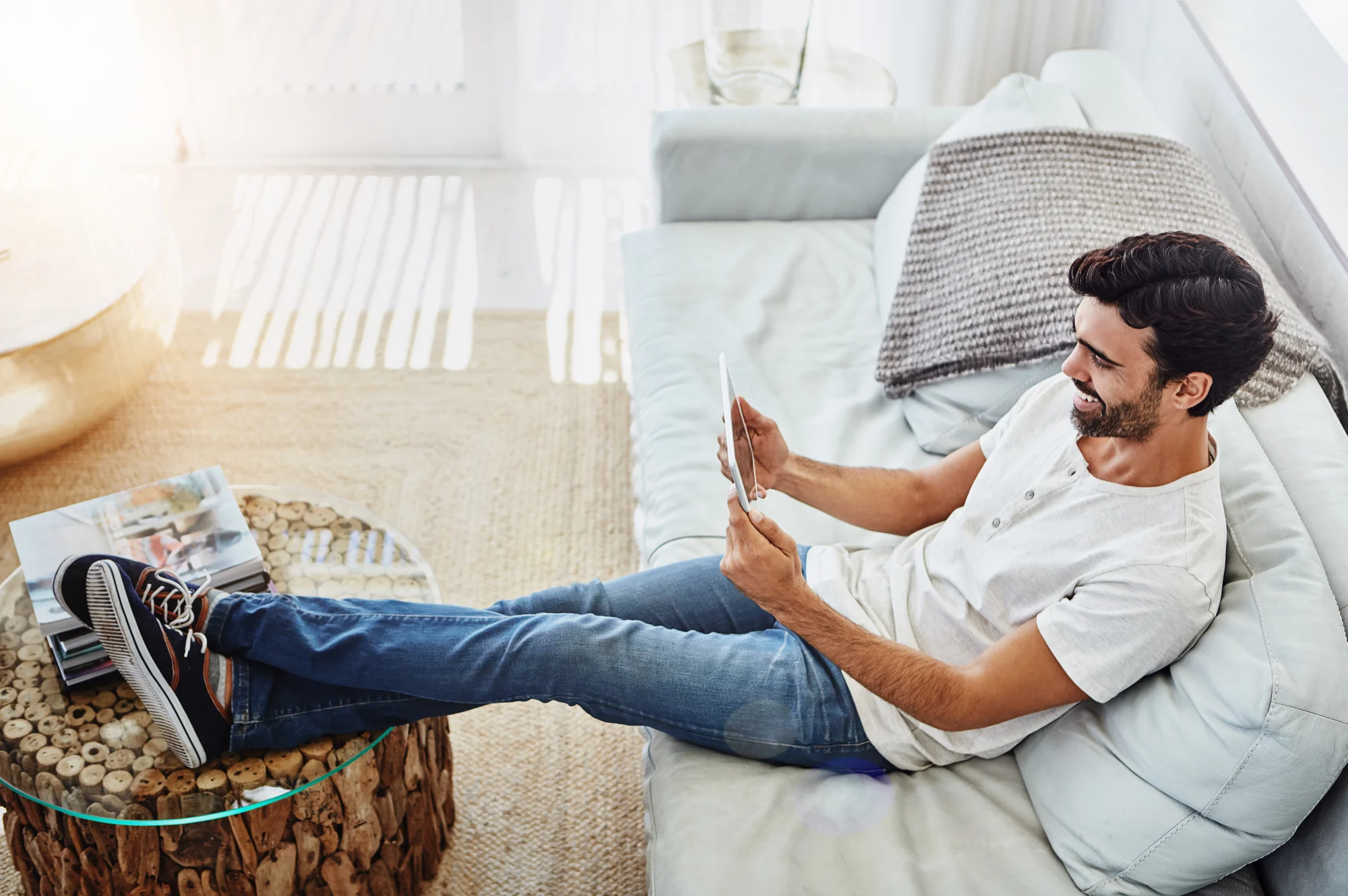 A man sitting comfortably on a gray house with his feet on a table while using his tablet.